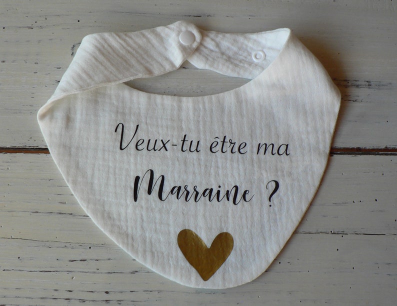 May include: White baby bib with the text "Veux-tu &ecirc;tre ma Marraine?" in black lettering and a gold heart.