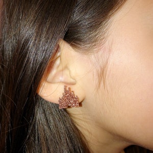May include: Rose gold castle stud earrings. The earrings are shaped like a castle with multiple towers and architectural details. The earrings are made of small, sparkling crystals. The earrings are worn on the ear of a person.