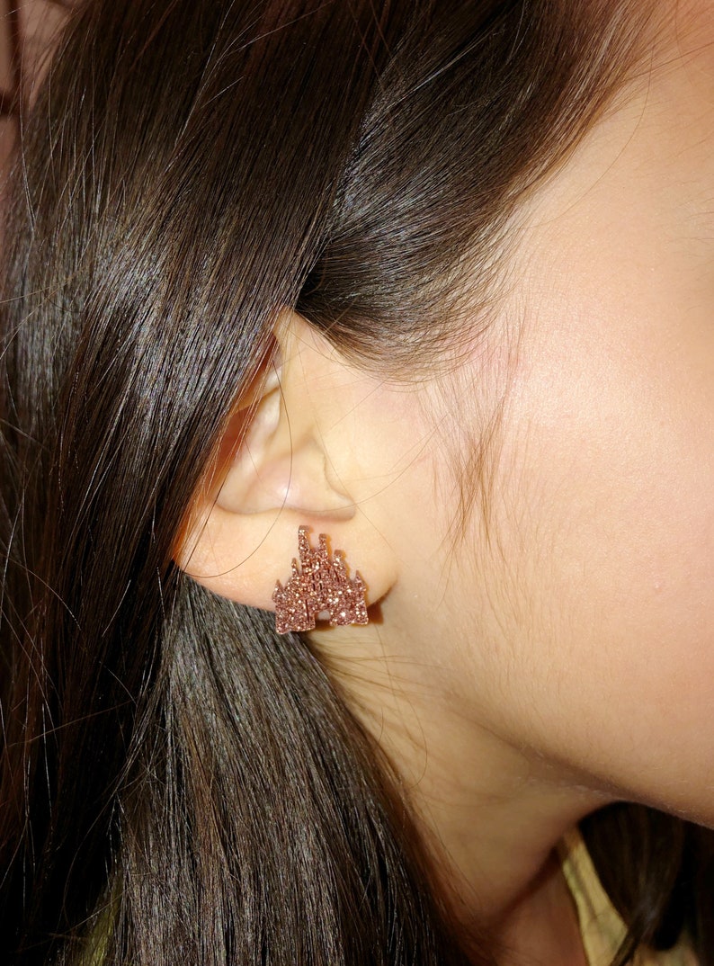 May include: Rose gold castle stud earrings. The earrings are shaped like a castle with multiple towers and architectural details. The earrings are made of small, sparkling crystals. The earrings are worn on the ear of a person.
