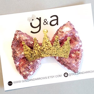 May include: A pink sparkly bow with a gold glitter crown on top. The bow is on a white card with the text "g&a" and "www.goldandarrows.etsy.com @goldandarrows".