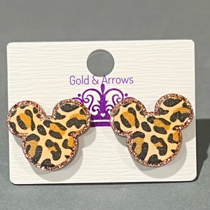 May include: A pair of Mickey Mouse-shaped earrings with a leopard print design. The earrings have a light brown background with black and orange spots. The edges are outlined with a glittery rose gold color. The earrings are displayed on a white card with the text "Gold & Arrows".