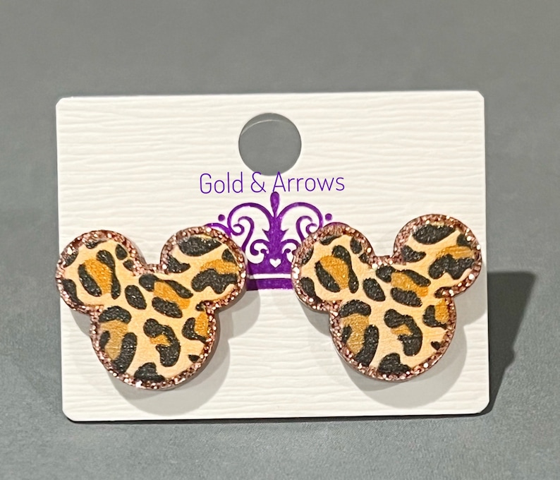May include: A pair of Mickey Mouse-shaped earrings with a leopard print design. The earrings have a light brown background with black and orange spots. The edges are outlined with a glittery rose gold color. The earrings are displayed on a white card with the text "Gold & Arrows".