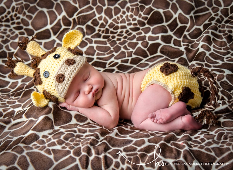 May include: A newborn baby wearing a yellow and brown crocheted giraffe hat and matching diaper cover, sleeping on a brown and white giraffe print blanket.