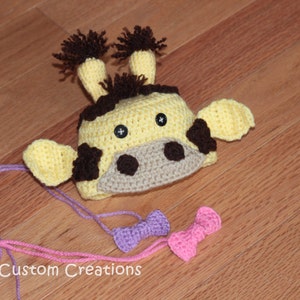 May include: A crocheted giraffe hat with brown ears and a yellow and brown face. The hat is on a wooden surface with two pink and purple bows.