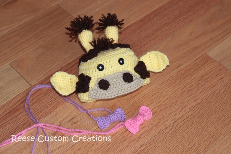 May include: A crocheted giraffe hat with brown ears and a yellow and brown face. The hat is on a wooden surface with two pink and purple bows.