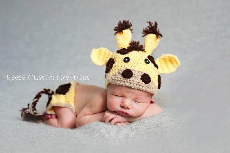 May include: A baby wearing a crocheted giraffe hat and matching diaper cover. The hat has brown spots and ears, and the diaper cover has a brown tail.