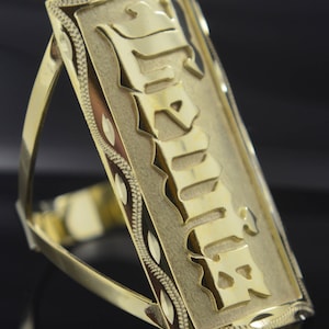 May include: A gold-colored ring with a rectangular band featuring the name "JULIA" in a bold, stylized font. The band has a decorative border with a repeating pattern.