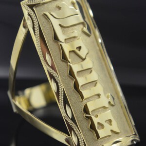 May include: A gold ring with a rectangular plaque featuring the word "Familia" in a bold, stylized font. The plaque has a textured, diamond-cut design around the edges.