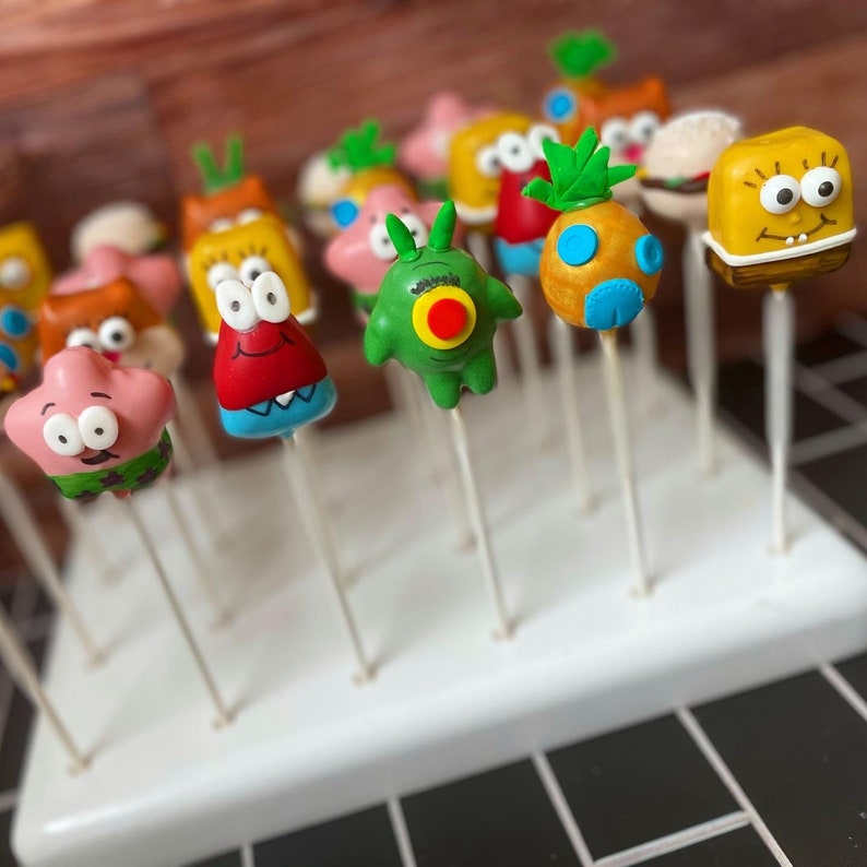 May include: A white cake pop stand with a variety of colorful cake pops decorated with cartoon characters. The cake pops are on sticks and are arranged in rows on the stand. The cake pops are decorated with different colors, including green, red, yellow, blue, and orange.