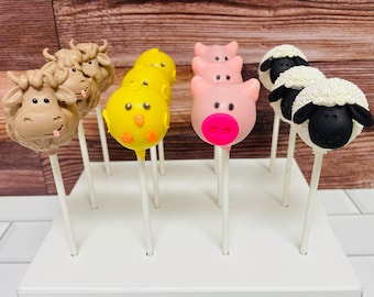 Può includere: Un supporto bianco per cake pops con una varietà di cake pops a tema animale. I cake pops sono decorati con mucche marroni, pulcini gialli, maiali rosa e pecore bianche.