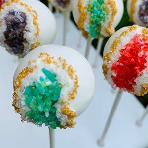 May include: White cake pops decorated with gold sprinkles and different colored sprinkles to resemble geodes. The cake pops are on white sticks.