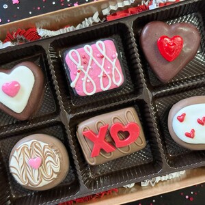 May include: A box of assorted chocolates, perfect for Valentine's Day. The chocolates are heart-shaped, square, and round, decorated with pink, white, and red icing. The box is brown with a clear plastic insert, and red and white paper shreds.