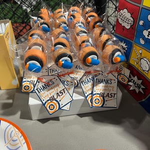 May include: A display of party favors, each individually wrapped. The favors are orange, black, and blue, resembling a design. Tags read "Thanks for making my party a BLAST." A comic book-themed gift bag is visible in the background.