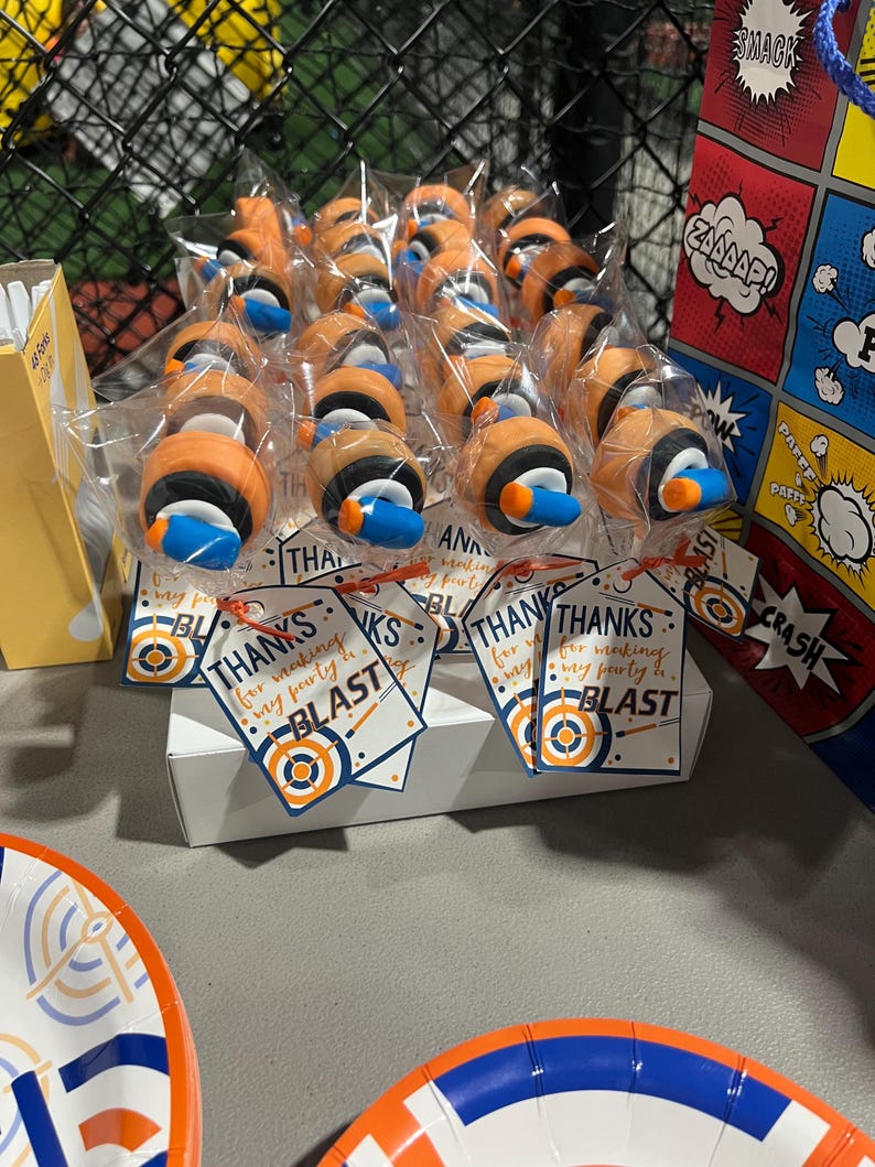 May include: A display of party favors, each individually wrapped. The favors are orange, black, and blue, resembling a design. Tags read "Thanks for making my party a BLAST." A comic book-themed gift bag is visible in the background.