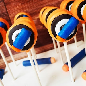 May include: Orange, black, white, and blue cake pops resembling toy darts. The pops have a cylindrical shape with orange frosting, black and white circular details, and a blue and orange accent. Several blue and orange dart-shaped candies are also present.