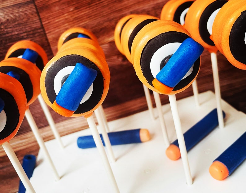 May include: Orange, black, white, and blue cake pops resembling toy darts. The pops have a cylindrical shape with orange frosting, black and white circular details, and a blue and orange accent. Several blue and orange dart-shaped candies are also present.