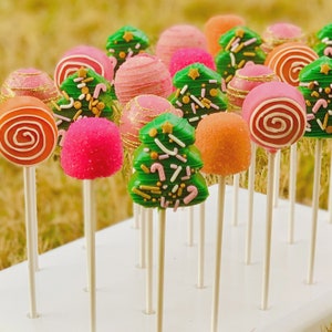 May include: A variety of Christmas-themed cake pops on sticks. The cake pops are decorated with green, pink, and orange frosting and sprinkles. Some of the cake pops are shaped like Christmas trees, while others are round.