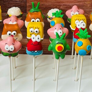 May include: A collection of 14 colorful cake pops decorated with cartoon characters. The cake pops are on white sticks and arranged on a white surface. The characters include SpongeBob SquarePants, Patrick Star, Squidward Tentacles, Sandy Cheeks, Plankton, and Mr. Krabs.