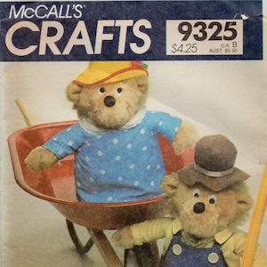Könnte beinhalten: McCall's Crafts 9325 Schnittmusterumschlag mit zwei Plüschbären. Ein Bär trägt einen gelben Hut und ein blaues Hemd mit weißen Punkten in einer roten Schubkarre. Der zweite Bär trägt einen braunen Hut und eine blaue Latzhose aus Jeansstoff.
