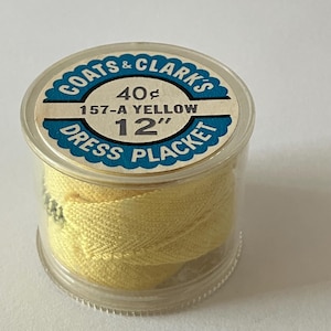 May include: A clear plastic container with a yellow fabric roll inside. The label on the container reads "Coats & Clark's Dress Placket 40¢ 157-A Yellow 12"