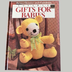 May include: A book titled "Gifts for Babies" with a crocheted yellow bear toy on the cover. The bear has a white face, black eyes, and a green and yellow striped bow. The book is on a wooden chair.