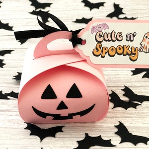 Puede incluir: Una caja de regalo rosa con tem&aacute;tica de Halloween con un asa negra y una etiqueta que dice "Cute n' Spooky". La etiqueta presenta una ilustraci&oacute;n de un murci&eacute;lago y un fantasma. Recortes de murci&eacute;lagos negros est&aacute;n esparcidos alrededor de la caja.