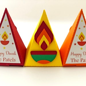 May include: Three colorful, triangular Diwali gift boxes. The boxes are red, yellow, and orange. Each box has a white label with the text "Happy Diwali The Patels" and a graphic of a lit diya and stars. The yellow box features a layered flame design.