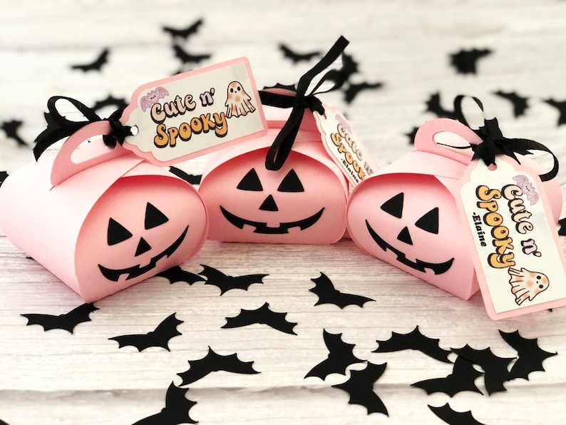 Puede incluir: Tres cajas de regalo rosas con tem&aacute;tica de Halloween con forma de calabaza, cada una con una cara de jack-o'-lantern negra. Cinta negra y etiquetas que dicen "Cute n' Spooky" con un dise&ntilde;o de fantasma y murci&eacute;lago. Recortes de murci&eacute;lagos negros esparcidos.