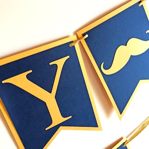 May include: A navy blue banner with gold accents. The banner features gold letters and a gold mustache cutout. The banner is strung together with gold string. The banner is likely for a party or celebration.