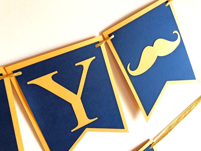 May include: A navy blue banner with gold accents. The banner features gold letters and a gold mustache cutout. The banner is strung together with gold string. The banner is likely for a party or celebration.