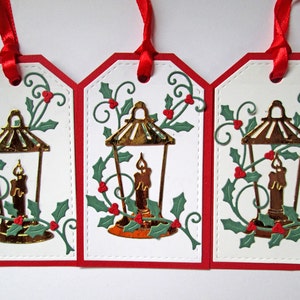 May include: Three Christmas gift tags with a white background, red border, and red ribbon. Each tag features a gold lantern with a lit candle, surrounded by green holly leaves and red berries.