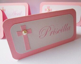 May include: Pink and white place card with a pink cross and a pearl. The name "Priscilla" is written in pink cursive.