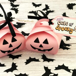 Puede incluir: Dos cajas de regalo rosas con tem&aacute;tica de Halloween con forma de calabaza, cada una con una cara de jack-o'-lantern negra. Asas de cinta negra y etiquetas que dicen "Cute n' Spooky" con una ilustraci&oacute;n de fantasma. Recortes de murci&eacute;lagos negros esparcidos.