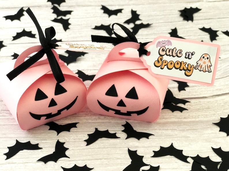 Puede incluir: Dos cajas de regalo rosas con tem&aacute;tica de Halloween con forma de calabaza, cada una con una cara de jack-o'-lantern negra. Asas de cinta negra y etiquetas que dicen "Cute n' Spooky" con una ilustraci&oacute;n de fantasma. Recortes de murci&eacute;lagos negros esparcidos.