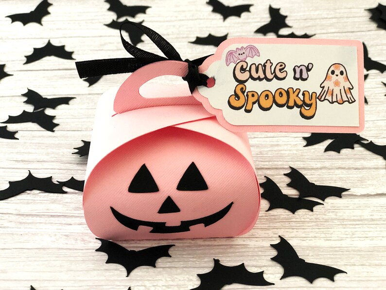 Puede incluir: Una caja de regalo rosa con tem&aacute;tica de Halloween con un asa negra y una etiqueta que dice "Cute n' Spooky". La etiqueta presenta una ilustraci&oacute;n de un murci&eacute;lago y un fantasma. Recortes de murci&eacute;lagos negros est&aacute;n esparcidos alrededor de la caja.