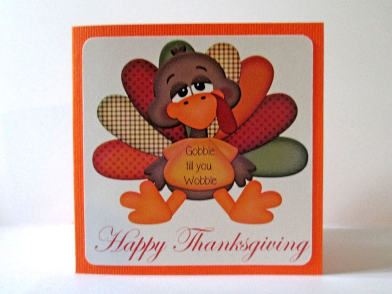 May include: Thanksgiving greeting card featuring a cartoon turkey with orange feet, beak, and wattle. The turkey has a brown body and colorful tail feathers. Text on the card reads "Gobble till you Wobble" and "Happy Thanksgiving."