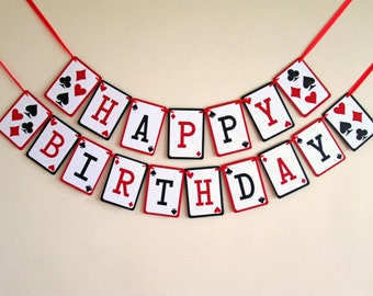 May include: A festive birthday banner made of playing card-shaped cutouts. Each card displays a letter spelling "HAPPY BIRTHDAY" in black and red, with card suit symbols. The banner is strung on a red ribbon.