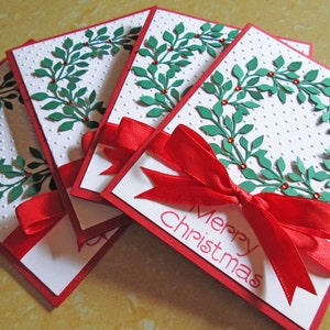 May include: Four Christmas cards with a white dotted background and a red border. Each card features a green leaf wreath design with red berries and a red ribbon. The words "Merry Christmas" are printed on the front.
