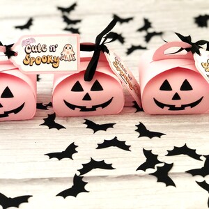 Puede incluir: Tres cajas de regalo rosas con tem&aacute;tica de Halloween con forma de calabaza, cada una con un corte de cara sonriente negro. Cintas negras y etiquetas que dicen "Cute n' Spooky" est&aacute;n adjuntas. Confeti de murci&eacute;lagos negros esparcido.