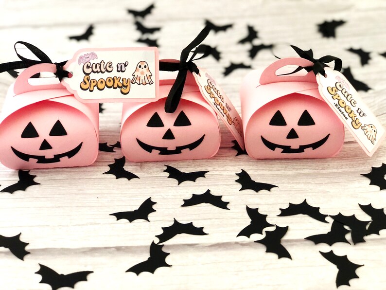 Puede incluir: Tres cajas de regalo rosas con tem&aacute;tica de Halloween con forma de calabaza, cada una con un corte de cara sonriente negro. Cintas negras y etiquetas que dicen "Cute n' Spooky" est&aacute;n adjuntas. Confeti de murci&eacute;lagos negros esparcido.