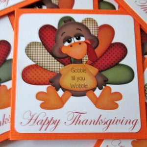 May include: Thanksgiving-themed square card with an orange border. The card features a cartoon turkey with a brown body, orange beak and feet, and colorful tail feathers. Text on the card reads "Gobble till you Wobble" and "Happy Thanksgiving".