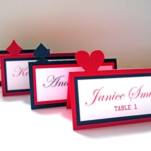 Peut inclure: Cartes de table rouges, noires et blanches avec des symboles de jeu de cartes. Chaque carte présente un nom en écriture cursive. La carte en forme de cœur est étiquetée "Janice Smith, TABLE 1."