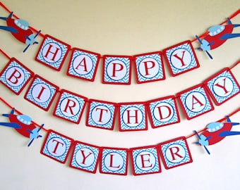Puede incluir: Bandera roja, blanca y azul con las palabras "Happy Birthday Tyler" en letras blancas sobre un patrón de chevron azul y blanco. La bandera está enhebrada con una cuerda roja y tiene aviones de papel rojos unidos.