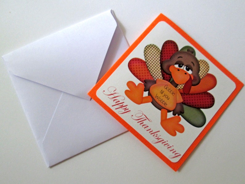 May include: Thanksgiving greeting card with a cartoon turkey illustration. The turkey has brown feathers, an orange beak and feet, and a red wattle. The card reads "Happy Thanksgiving" and "Gobble til you Wobble". A white envelope is next to the card.