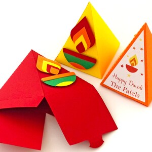 May include: Three colorful, triangular Diwali-themed gift boxes. One is red and open, another is yellow with a flame design, and the third is orange with the text "Happy Diwali The Patels".