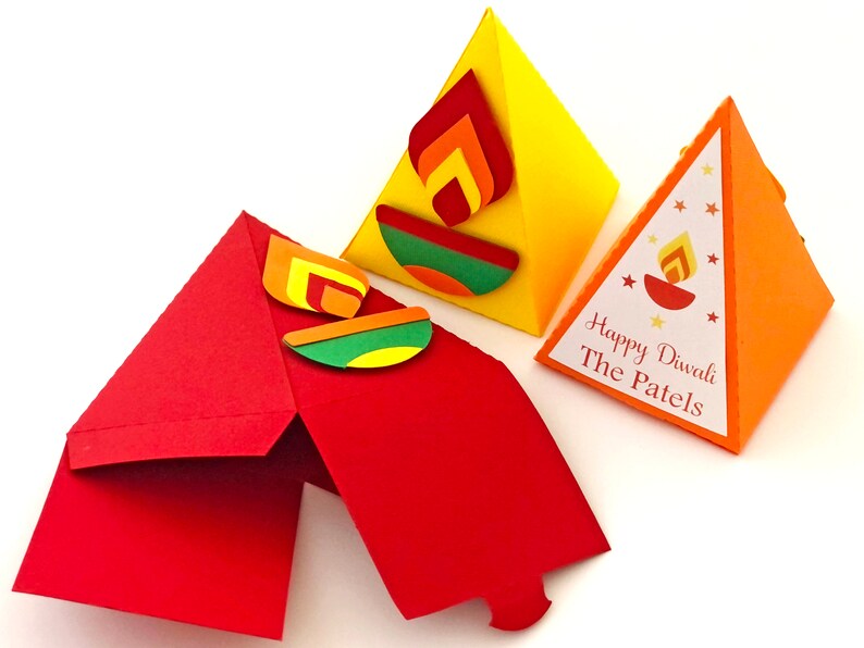 May include: Three colorful, triangular Diwali-themed gift boxes. One is red and open, another is yellow with a flame design, and the third is orange with the text "Happy Diwali The Patels".