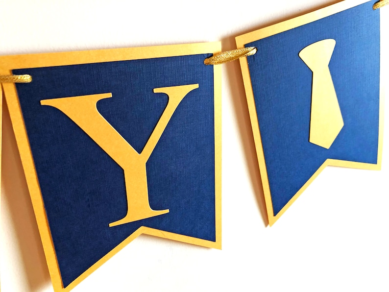 May include: A banner with navy blue pennants and gold accents. The pennants are strung together with gold cord. One pennant has a gold letter "Y", and another has a gold tie shape. The banner is likely for a party or celebration.
