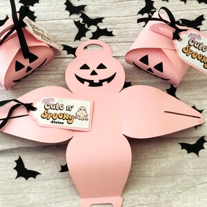 Puede incluir: Cajas de regalo rosas con tem&aacute;tica de Halloween con caras de calabaza talladas y lazos negros. Cada caja tiene una etiqueta que dice "Cute n' Spooky" con una ilustraci&oacute;n de fantasma. Recortes de murci&eacute;lagos negros esparcidos.