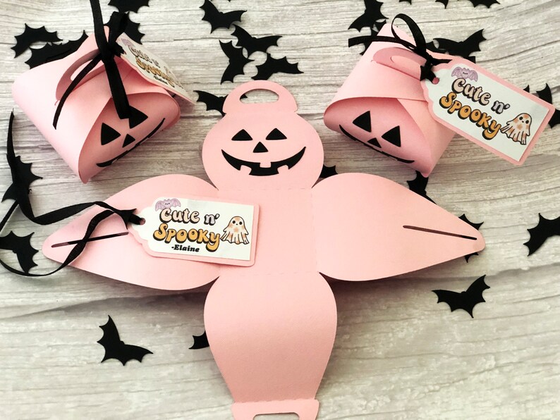 Puede incluir: Cajas de regalo rosas con tem&aacute;tica de Halloween con caras de calabaza talladas y lazos negros. Cada caja tiene una etiqueta que dice "Cute n' Spooky" con una ilustraci&oacute;n de fantasma. Recortes de murci&eacute;lagos negros esparcidos.