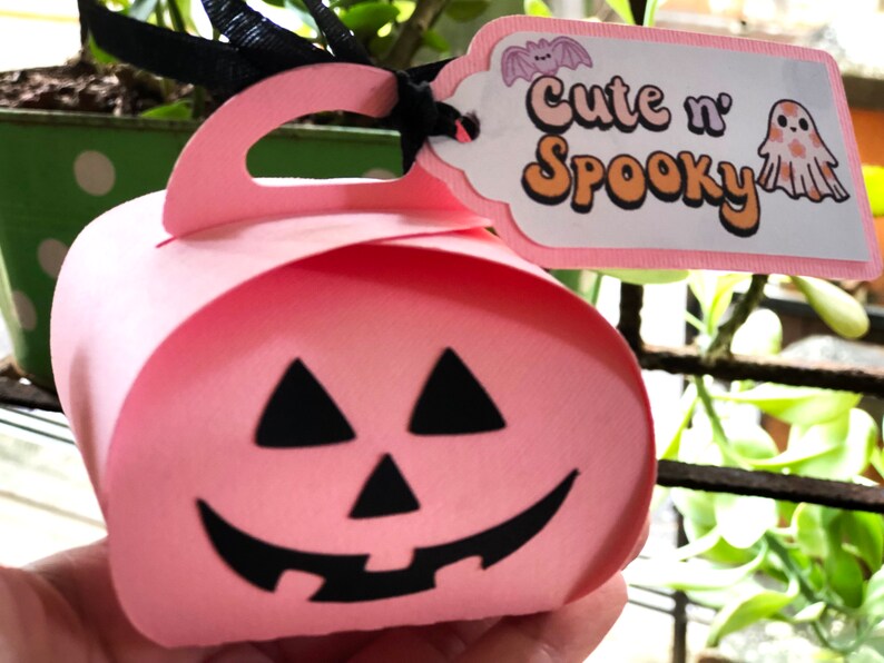 Puede incluir: Una caja de regalo rosa con tem&aacute;tica de Halloween con forma de calabaza con una cara negra. La caja tiene un asa y una etiqueta que dice "Cute n' Spooky" con una ilustraci&oacute;n de murci&eacute;lago y fantasma. Se adjunta una cinta negra.